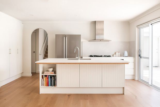 Bright, contemporary open-plan kitchen with a large central island featuring white cabinetry, vertical timber-style panelling, an integrated sink and seating overhang. The room has warm timber flooring, white walls and cabinetry, stainless-steel refrigerator and rangehood, a tiled or light splashback, and expansive sliding glass doors opening to the exterior. An arched doorway leads toward a stairway or hallway.