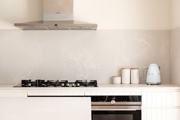 Bright, minimalist kitchen focusing on a gas cooktop, stainless-steel rangehood, pale stone-look benchtop, tiled splashback, and integrated stainless-steel oven. White cabinetry with simple vertical detailing and a restrained neutral palette create a clean contemporary look.