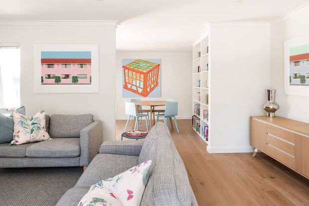 Bright open-plan living and dining area with white walls, traditional ceiling cornices, light timber flooring, grey upholstered sofas, and a compact dining setting. Built-in white shelving separates the living zone from the dining area, while a mid-century-style timber sideboard sits against the right wall. Colourful framed artwork and floral cushions add playful accents.