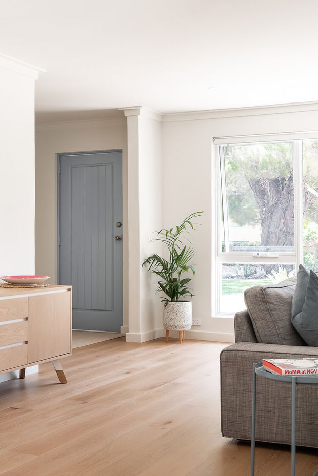 Bright, refined living room with a grey upholstered sofa beside a large window, a light timber mid-century-style sideboard, pale timber flooring, and a grey paneled entry door. Cream walls, simple ceiling cornices, and a potted palm create an airy contemporary interior with subtle traditional detailing.