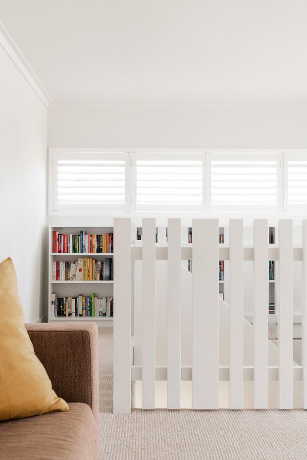 Bright, predominantly white upper-level interior featuring a brown upholstered sofa in the foreground, a white vertical-slat balustrade, built-in bookshelves filled with books, plantation shutters, and a pale textured carpet. The image appears to look across a living area toward an open stair landing or stairwell, with clean contemporary detailing and abundant daylight.