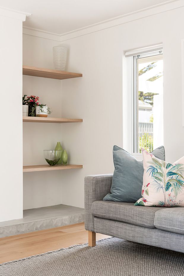 Bright, refined living room corner with a grey upholstered sofa, blue and botanical-patterned cushions, a textured grey rug, pale timber flooring, and built-in recessed shelving with light timber shelves and decorative ceramics and plants. White walls, ceiling trim, and a large window create an airy, calm atmosphere.