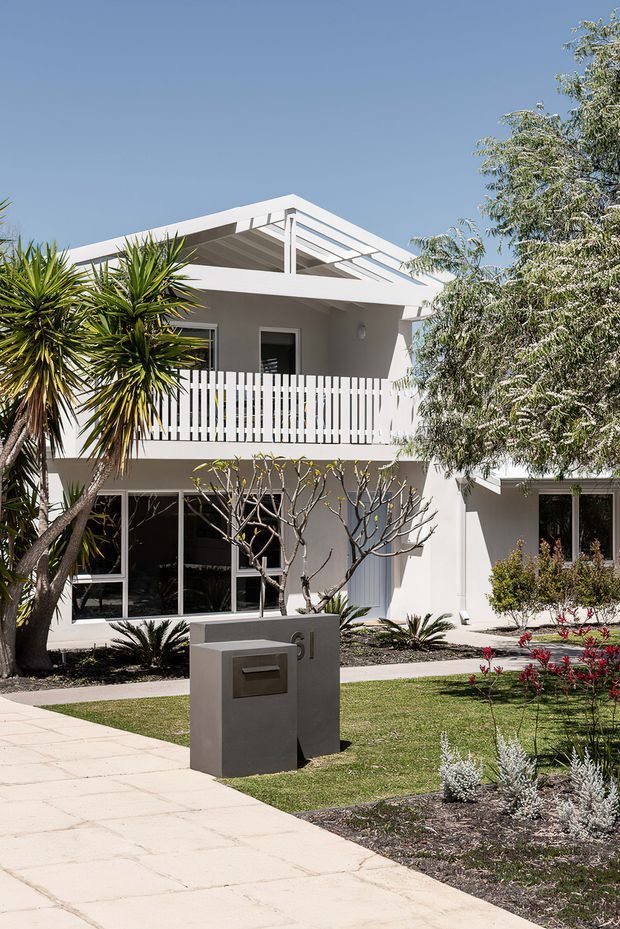 Bright daytime photograph of a two-storey white residential house viewed from the front garden. The home has a prominent gabled roof with exposed white structural rafters, large glazed openings, an upper-level balcony with vertical white balustrade, and a landscaped front yard with palms, low planting, lawn, paved walkway, and a rendered mailbox structure.