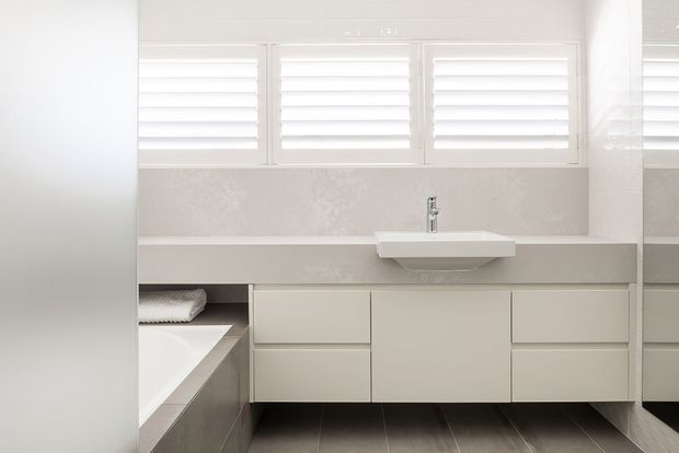Bright, contemporary bathroom with a long white floating vanity, integrated drawers and cabinetry, a wall-mounted rectangular basin with chrome tapware, and a partially visible bathtub. White plantation shutters span the upper wall, while grey textured wall finish and dark grey floor tiles add contrast. A large mirror is visible at the right edge.