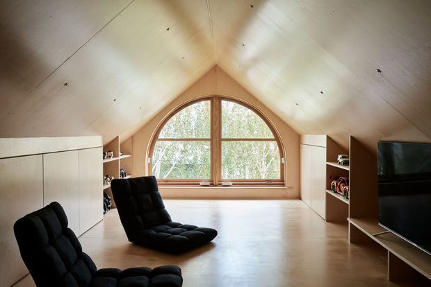 A warm attic-style living space with a steeply pitched vaulted ceiling, pale timber or wood-finished surfaces, built-in storage along both side walls, and a large arched timber-framed window overlooking trees. Two black cushioned lounge chairs and a television create a relaxed media or sitting area.