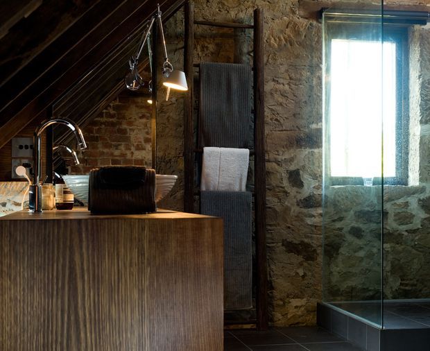 Rustic bathroom interior with exposed rough stone and brick walls, a dark timber vanity with integrated basin and chrome tapware, a glass-enclosed shower, dark tiled flooring, and a ladder-style towel rack. Warm pendant lighting and timber roof structure create an intimate, moody atmosphere.