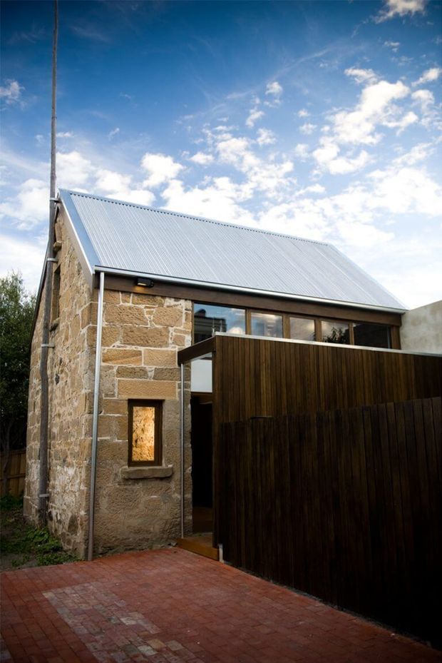 Exterior view of a compact renovated stone building with a steeply pitched corrugated metal gable roof. The facade combines exposed warm sandstone masonry, dark timber privacy fencing and cladding, and a long band of glazing beneath the roof edge. A red brick-paved walkway occupies the foreground, with a small wall-mounted exterior light and a tall slender pole visible against a bright blue sky.