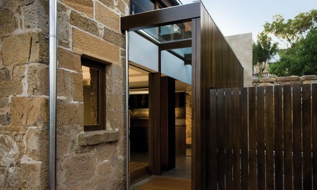Exterior view of a contemporary addition integrated with an older rough-hewn stone building. The addition features dark timber cladding, a tall glazed entry with exposed structural framing, and a matching vertical timber privacy fence. Warm interior lighting is visible through the doorway, while the original stone wall and small timber-framed window remain prominent.