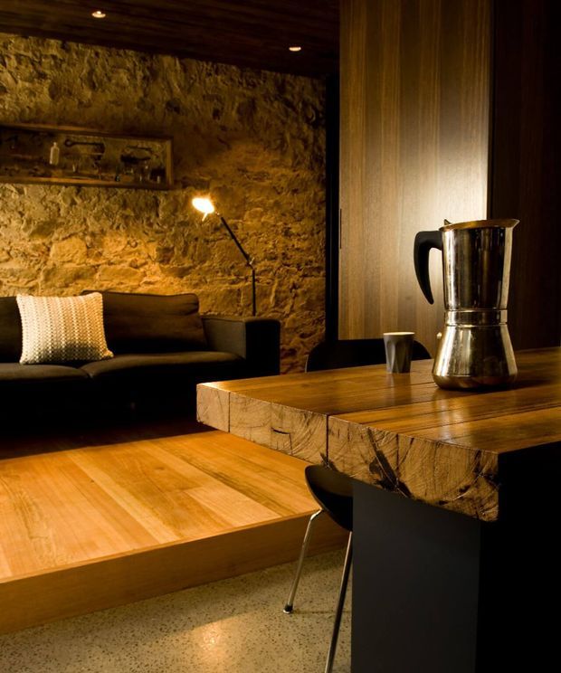 Warm, moody interior combining a rustic stone feature wall and dark timber ceiling with contemporary timber furniture. A low black sofa sits beneath a wall shelf and floor lamp, while a substantial timber dining or work table with a metal coffee pot and stools occupies the foreground. The room has polished speckled concrete flooring and integrated recessed lighting, creating a refined rustic-industrial atmosphere.