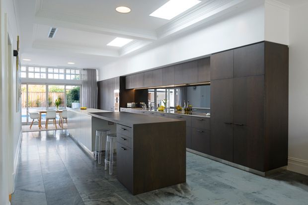 A bright, spacious open-plan kitchen with a large dark timber island, extensive dark cabinetry, mirrored splashback, and integrated appliances. The kitchen opens to a dining area and glazed outdoor connection at the rear. White walls and coffered detailing, skylights, and polished grey stone-like flooring create a refined contemporary interior.