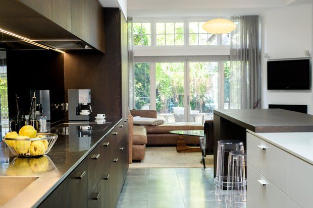 Open-plan contemporary kitchen and living area. The kitchen features dark timber cabinetry, a dark stone benchtop, black splashback and integrated lighting, with a contrasting white island and stools. Beyond it is a compact living area with a sofa, coffee table, large multi-pane windows and sheer curtains. The space is bright with a warm, refined palette and strong indoor–outdoor visual connection.