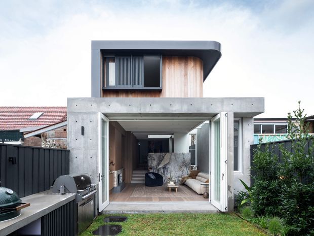 Rear exterior view of a contemporary residential extension with a projecting upper level, curved dark roof edge, concrete framing, timber-look wall cladding, and large open bi-fold doors leading into a living area. A covered outdoor cooking area and garden sit in the foreground, with neighbouring roof forms visible behind.