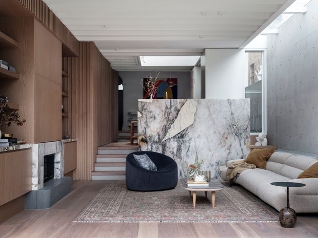 A refined contemporary living room with warm timber built-in joinery and shelving along one wall, a dark fireplace surround, pale timber flooring, a large patterned area rug, and a sculptural dark lounge chair. A light stone-clad partition separates the seating area from a raised dining or circulation zone. The room features a white exposed-beam ceiling with skylights, polished concrete walls, a large curved light sofa, and restrained neutral furnishings.