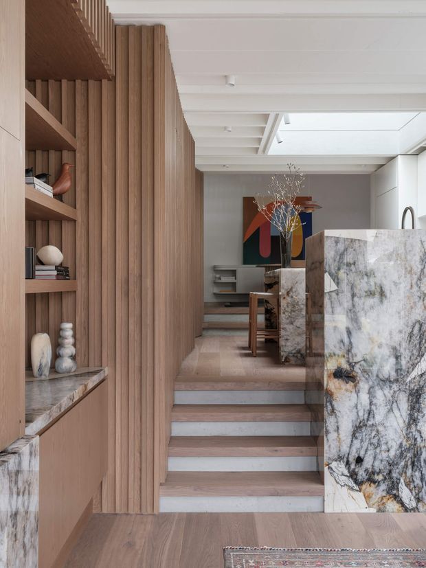 A refined open-plan residential interior viewed from a lower level, featuring a timber-lined stair and vertical timber slat partition, built-in shelving and cabinetry, a dramatic veined stone kitchen island, and a bright white coffered ceiling with a skylight. The space combines warm natural materials with contemporary detailing and a restrained palette.