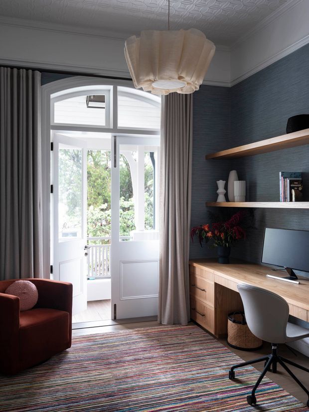 A refined home office with a built-in timber desk and drawers, wall-mounted open shelving, a computer workstation, and a sculptural upholstered office chair. The room features a dark textured blue-grey wall, white decorative pressed-metal ceiling and ornate trim, tall glazed French doors opening to a balcony, full-height grey curtains, and a colourful striped area rug. A burgundy armchair and pendant light add warmth and character.