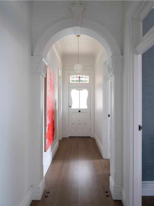 A narrow, bright residential entry hall viewed toward a white front door. The hallway has warm timber flooring, white walls and ornate moulded architraves, a large decorative arch with carved capitals, a hanging globe pendant, and a small stained-glass transom above the door. A vivid red artwork is mounted on the left wall, while an adjacent room is partially visible through the opening on the right.