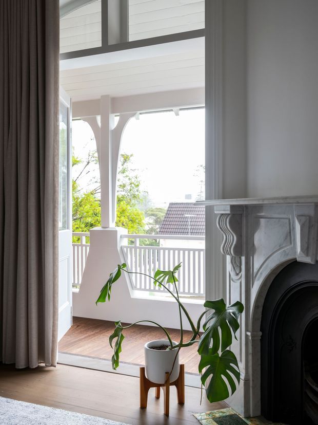 Bright interior view toward a covered balcony or veranda, with a decorative marble fireplace in the foreground, tall glazed doors, timber flooring, and a potted monstera. The veranda features white balustrading, timber decking, and distinctive curved decorative supports, with leafy suburban surroundings beyond.