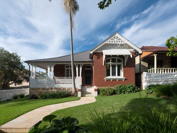 Street-facing view of a traditional single-storey brick bungalow with a prominent front gable, decorative timber fretwork, verandah, white balustrades, tiled roof sections, and a landscaped front garden with a curved paved path.