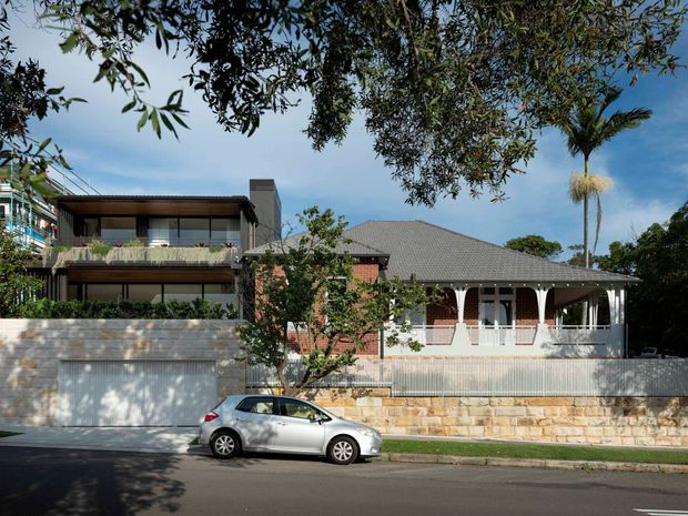 Street-facing view of a residential property combining a contemporary multi-level addition with an older single-storey house. The home has dark horizontal cladding and glazing, a broad low-pitched tiled roof, red-brick walls, white decorative verandah arches, stone retaining walls, and a rendered garage frontage. Mature trees and a parked car are visible in the foreground.