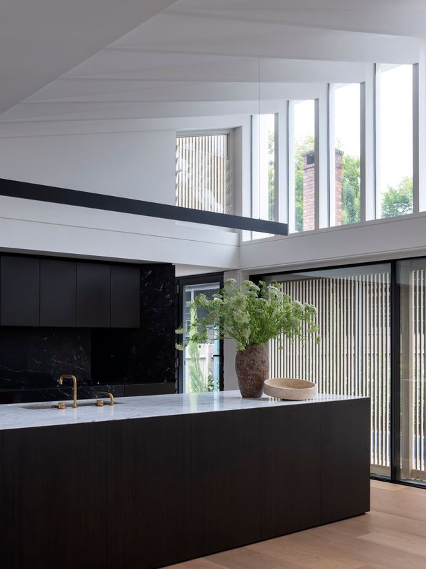 A refined contemporary kitchen with a large dark island, pale stone benchtop, matte black cabinetry and dark marble-look splashback. White structural beams and a high glazed wall create an airy, double-height feel, with vertical blinds and timber flooring adding warmth. A sculptural vase and greenery sit on the island.