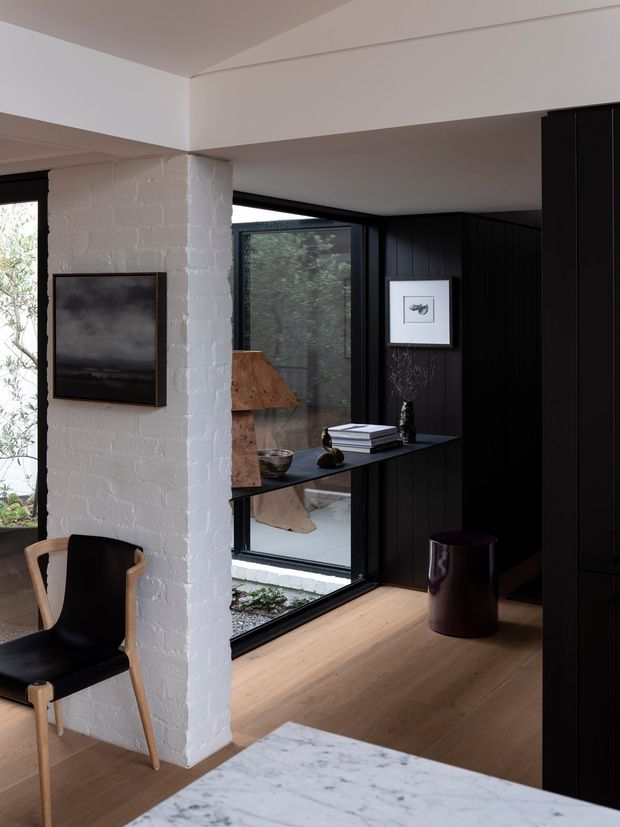 Interior view of a refined contemporary living and dining area with a white-painted exposed brick structural wall, dark vertical timber-panelled joinery, black-framed floor-to-ceiling glazing, and warm timber flooring. A black and timber armchair, console styling, artwork, and sculptural lighting create a moody, minimalist composition. A marble-topped surface is visible in the foreground, with greenery seen through the glazing.