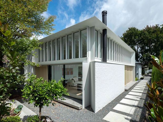 Exterior view of a compact two-storey contemporary house set within a landscaped garden. The white rectilinear volume has extensive narrow vertical glazing on the upper level, a projecting flat roof, a white painted brick ground-floor wall, dark metal chimney, and a paved stepping-stone path through a gravel forecourt. Interior living furnishings are visible through the ground-floor glazing.