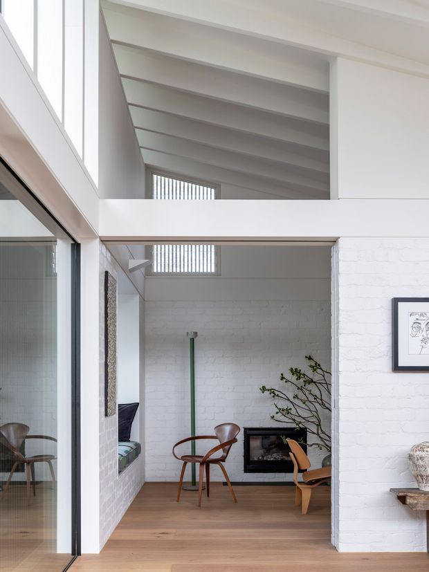 Bright double-height interior with a loft-like open mezzanine and exposed white-painted structural roof framing. White-painted brick walls, pale timber flooring, large dark-framed glazing, high clerestory windows, and a compact seating area with mid-century timber chairs surround a black fireplace. Built-in shelving and an open void create a strong vertical connection between levels.