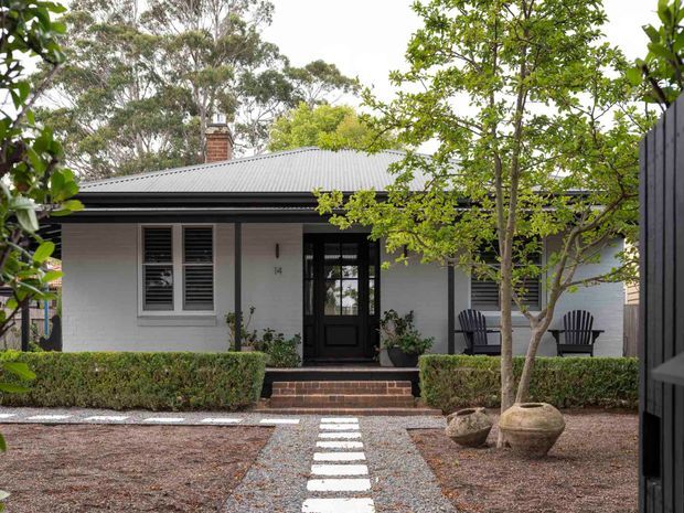 Front exterior view of a single-storey rendered brick or masonry home with a low-pitched corrugated metal roof, dark trim, black entry door, and shuttered window. A formal landscaped entry garden includes clipped hedges, gravel, stepping stones, mature trees, potted plants, and a small covered porch with outdoor chairs.