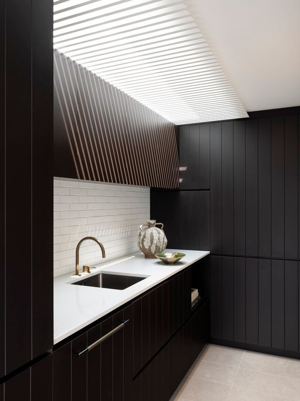 A refined compact kitchen with dark vertically panelled cabinetry, a white engineered-stone benchtop, integrated sink and brass-toned curved tapware. White subway tile forms the splashback, while a dramatic slatted timber feature extends across the ceiling and upper wall, creating a strong linear architectural detail. The room is brightened by a large overhead skylight or linear clerestory-like opening, with pale stone-look flooring and restrained decorative styling.