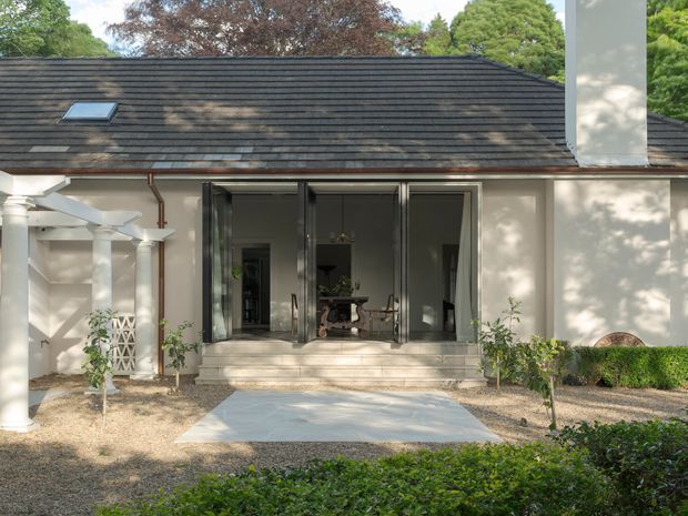 A single-storey residential rear facade opening onto a landscaped courtyard or garden. The pale rendered exterior has a dark pitched tiled roof with a skylight, tall black-framed glazed folding or sliding doors, and a projecting covered entry or pergola supported by white columns at left. Steps lead to a paved path through gravel and clipped planting. A dining area is visible through the open glazing.