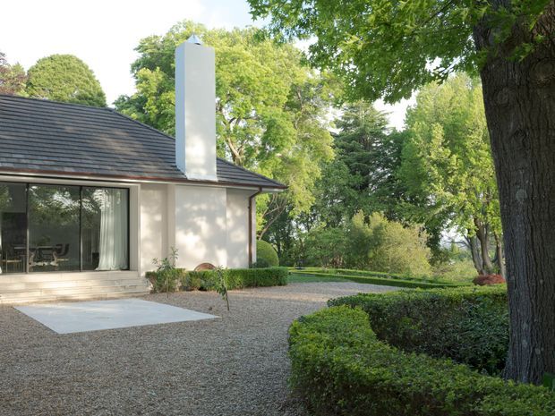 Exterior view of a refined, low-profile residence set within a mature landscaped garden. The house has a dark pitched roof, pale rendered walls, a tall white chimney, and large glazed sliding doors opening onto a gravel courtyard with clipped hedges and established trees.