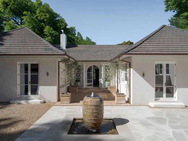 A symmetrical single-storey courtyard facade with pale rendered walls, dark shingle-clad hipped roofs, French doors, arched openings, and climbing greenery. A central barrel-style water fountain sits in a paved stone forecourt, creating a tranquil Mediterranean-inspired outdoor setting.