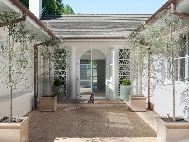 A bright, refined courtyard entry framed by pale rendered walls and pitched tiled roof forms. The central arched doorway opens into a light-filled interior, flanked by decorative white lattice screens and tall glazed windows. Small olive trees in large rectangular planters line the herringbone brick-paved courtyard, creating a formal Mediterranean-inspired approach.