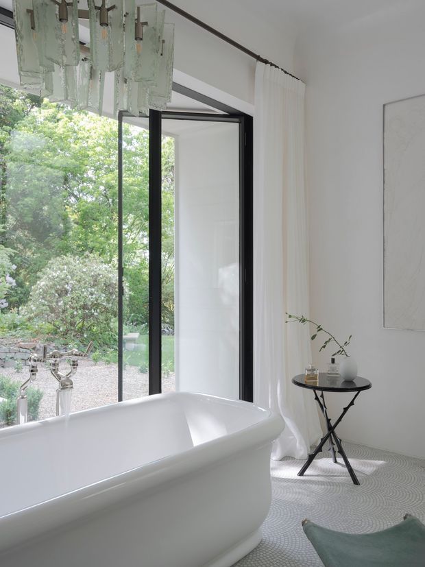 Bright, refined bathroom featuring a freestanding white bathtub beside expansive black-framed glazing, sheer white curtains, and a lush garden view. A sculptural translucent pendant hangs overhead, while a small black side table with minimal decor and a pale artwork add understated elegance. The floor has a subtle patterned finish and the room has a calm, light-filled atmosphere.