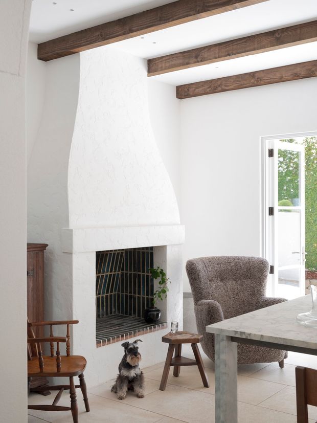 Bright, airy living room with a large white textured masonry fireplace, exposed dark timber ceiling beams, pale tiled flooring, and an open glazed door to the exterior. A sculptural upholstered armchair, timber furniture, and a marble-look table complete the warm, rustic-contemporary interior. A dog is present near the fireplace.