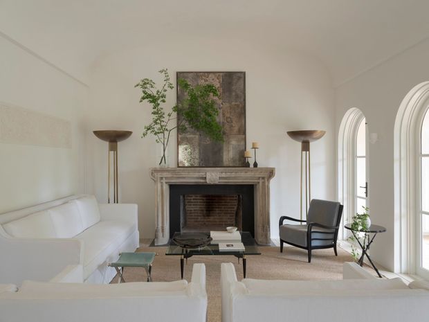 Refined living room with a soft neutral palette, centered classical fireplace, white upholstered sofas, a dark-framed armchair, slender floor lamps, and tall arched glazed doors. A weathered artwork and potted greenery add understated natural texture. The room feels bright, calm, and elegant, with a traditional architectural shell styled in a restrained contemporary-classic manner.