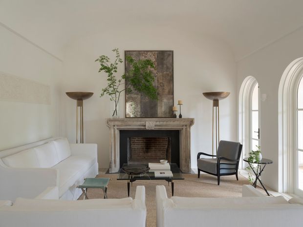 A bright, refined living room centered on a traditional stone or plaster fireplace with exposed brick firebox. White sofas face the fireplace, accompanied by a dark upholstered armchair, slender floor lamps, a glass coffee table, and a large abstract artwork. Tall arched glazed doors or windows line the right side, while neutral walls and carpeting create an airy, calm atmosphere.