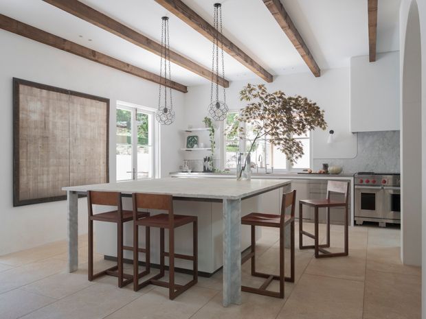 A bright, open-plan kitchen and dining area with a large pale stone island, four dark timber bar stools, white cabinetry, stainless-steel appliances, and a subtle grey textured backsplash. Exposed dark timber ceiling beams, pendant lights, arched wall detailing, and large windows give the space a warm contemporary character.