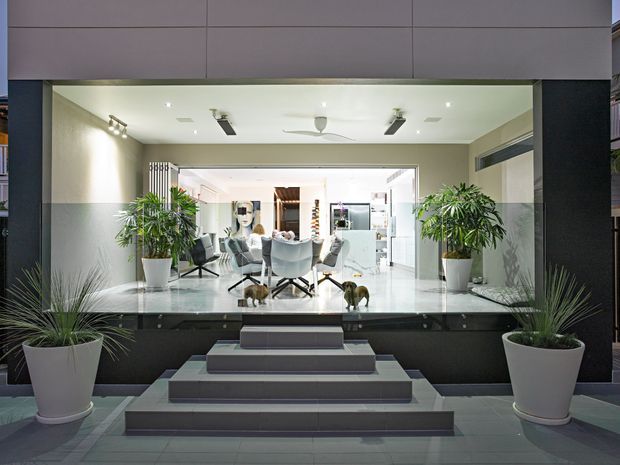 A contemporary residential entry facade viewed from outside, with broad dark and grey cladding framing a fully glazed recessed entrance. Wide tiled steps lead to a shallow landing, flanked by large white planters with palms and grasses. Through the glazing, a bright open-plan interior with a dining table, upholstered chairs, kitchen cabinetry and pendant lighting is visible. The composition is symmetrical and polished, with strong indoor-outdoor visual connection.