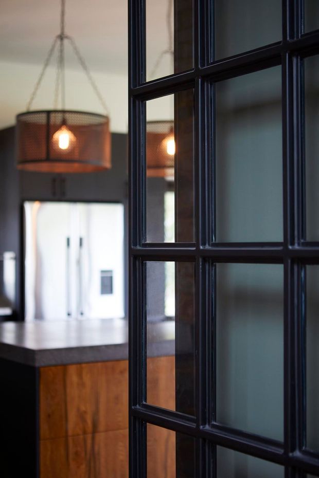 A close-up interior view through a black-framed, multi-pane glazed partition or door, looking toward a kitchen. The kitchen features warm timber cabinetry, a dark benchtop, pale walls, and a hanging pendant light with a perforated metal shade. The strong black framing creates an industrial-inspired architectural detail.