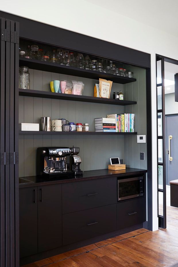 A compact built-in coffee and beverage station with dark cabinetry, open black shelving, and a charcoal vertical-panelled backsplash. The shelves display glassware, mugs, pantry items, books, and accessories, while a professional espresso machine sits on the dark benchtop. Warm timber flooring contrasts with the dark joinery, and a glazed black-framed doorway opens to an adjacent room.