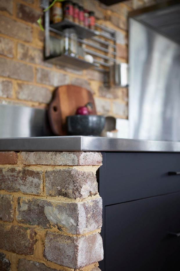 Close-up detail of a rustic industrial kitchen featuring exposed reclaimed brickwork, a dark matte kitchen island or cabinetry, a stainless-steel benchtop, and open metal shelving displaying jars and kitchen items. Warm natural materials contrast with dark joinery and cool metal surfaces.