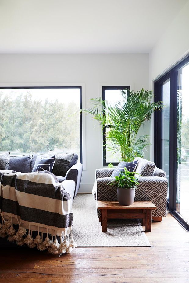 Bright, airy living room with large black-framed windows and floor-to-ceiling glazing overlooking leafy greenery. The room features a grey sofa draped with a black-and-white fringed throw, a patterned upholstered armchair, a small timber coffee table, indoor palms and plants, a pale textured rug, white walls, and warm timber flooring.