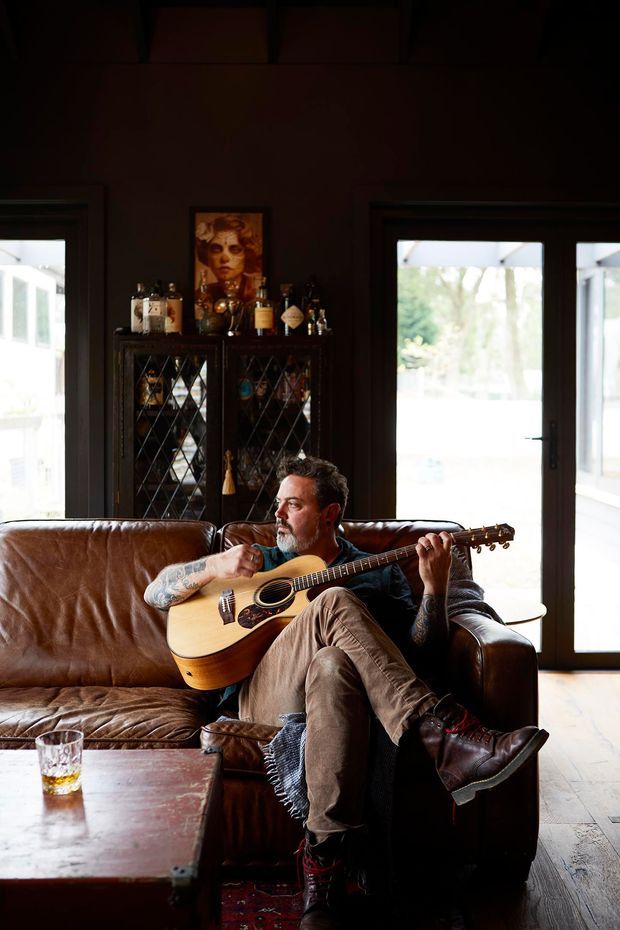A warmly lit living room with dark painted walls, exposed timber ceiling beams, large dark-framed glazed doors and windows, and wide timber flooring. A brown leather sofa faces the camera, with a man playing an acoustic guitar. Behind the sofa is a glass-fronted drinks cabinet and a framed portrait, while a timber coffee table and patterned rug complete the relaxed, characterful interior.