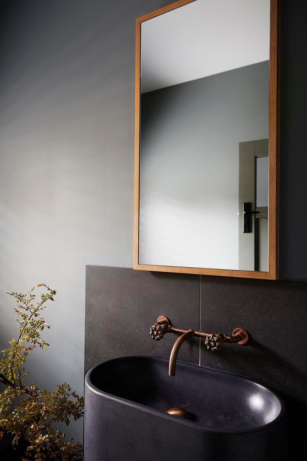 A moody bathroom vignette featuring a large black vessel basin with a wall-mounted antique bronze tap set, dark textured wall panels, and a rectangular timber-framed mirror. A leafy plant adds an organic accent beside the basin.