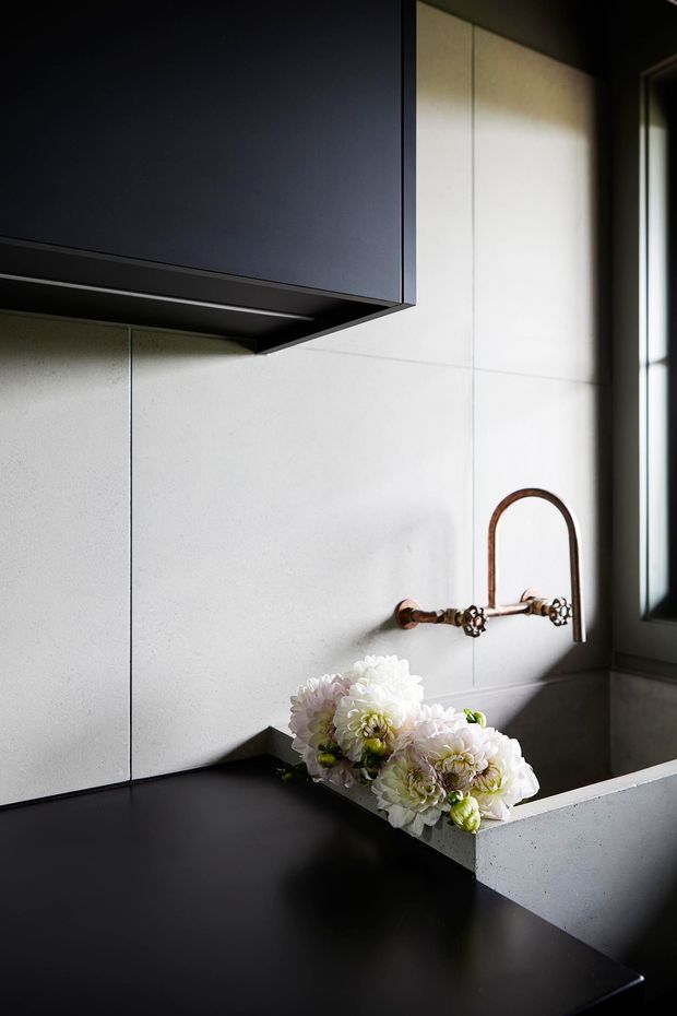 A close-up of a refined bathroom vanity area featuring a matte black benchtop and dark overhead cabinetry, pale large-format wall tiles, a sculptural antique-brass wall-mounted tap, and a white inset basin partially visible at the lower right. Natural light enters through a dark-framed window, while a floral arrangement rests beside the basin.
