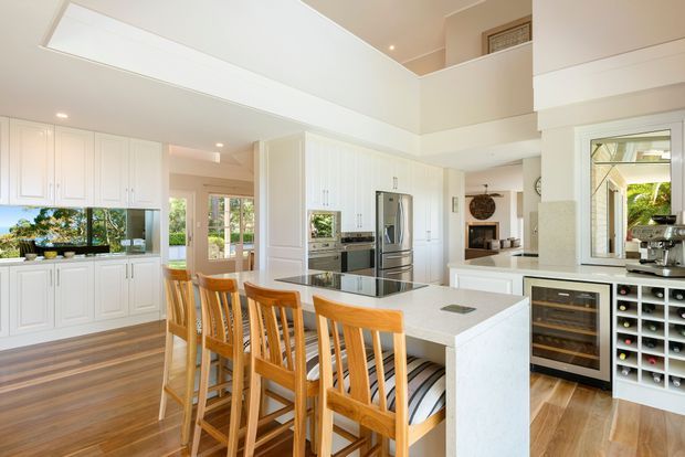Bright, spacious open-plan kitchen with extensive white paneled cabinetry, a large white island with timber bar stools, stainless-steel appliances, integrated wine storage, timber flooring, and a double-height ceiling with clerestory-style glazing. A living area and upper-level opening are visible beyond the kitchen.