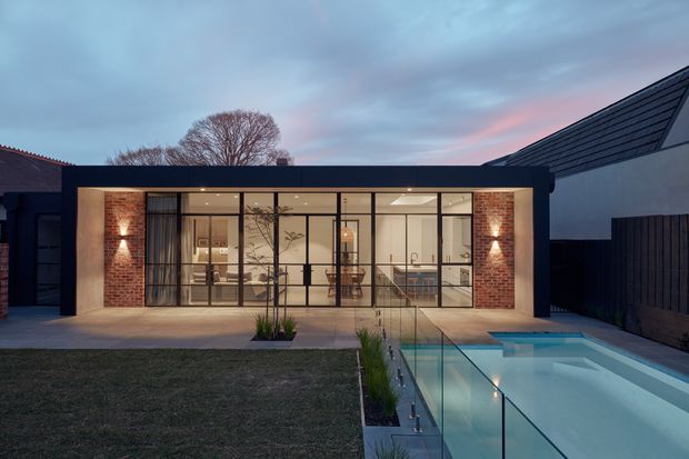 A contemporary single-storey residential extension viewed from the rear garden at dusk. The pavilion-like addition has a flat roof, extensive black-framed floor-to-ceiling glazing, exposed red-brick walls, and warm interior lighting. A glazed swimming pool and narrow planted strip run alongside the building, with dark fencing and neighbouring roof forms framing the scene.
