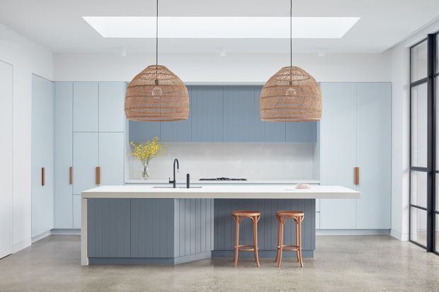 Bright contemporary kitchen with a large island, pale blue-grey flat-panel cabinetry, white benchtop, integrated cooktop and sink, polished concrete floor, black-framed glazing, and two large woven pendant lights. The clean, open layout has a skylight or ceiling lightwell above the island and timber bar stools at the seating side.