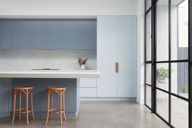 A bright, refined contemporary kitchen with a compact breakfast island, two timber stools, blue-grey vertically grooved cabinetry, white overhead and full-height cupboards, and a pale benchtop. Large black-framed glazed doors or windows line the right side, bringing in abundant daylight and providing views to a small planted outdoor area. The polished concrete or terrazzo-like floor and restrained palette create a clean, minimalist atmosphere.